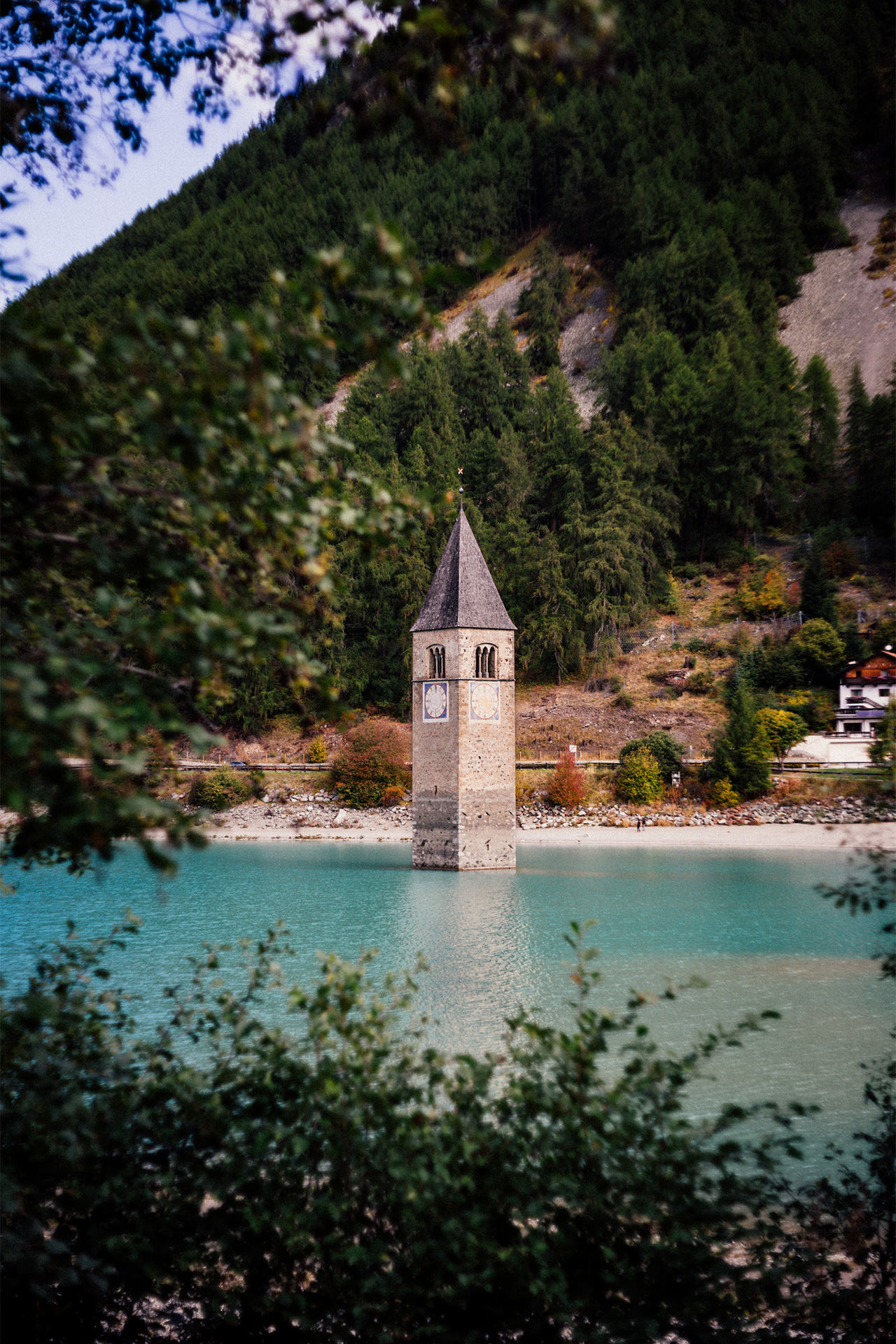Ein Kirchturm der in einem See steht am fuße eines Berges. Im Vordergrund sind verschwommene Äste von Bäumen.